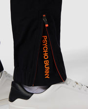 Close-up of the lower leg of black MENS MADISON COMMUTER PANT - B6P395Z1WB by Psycho Bunny, showcasing a zipper detail with the text "psycho bunny" in orange along the seam, paired with white shoes on a white background.