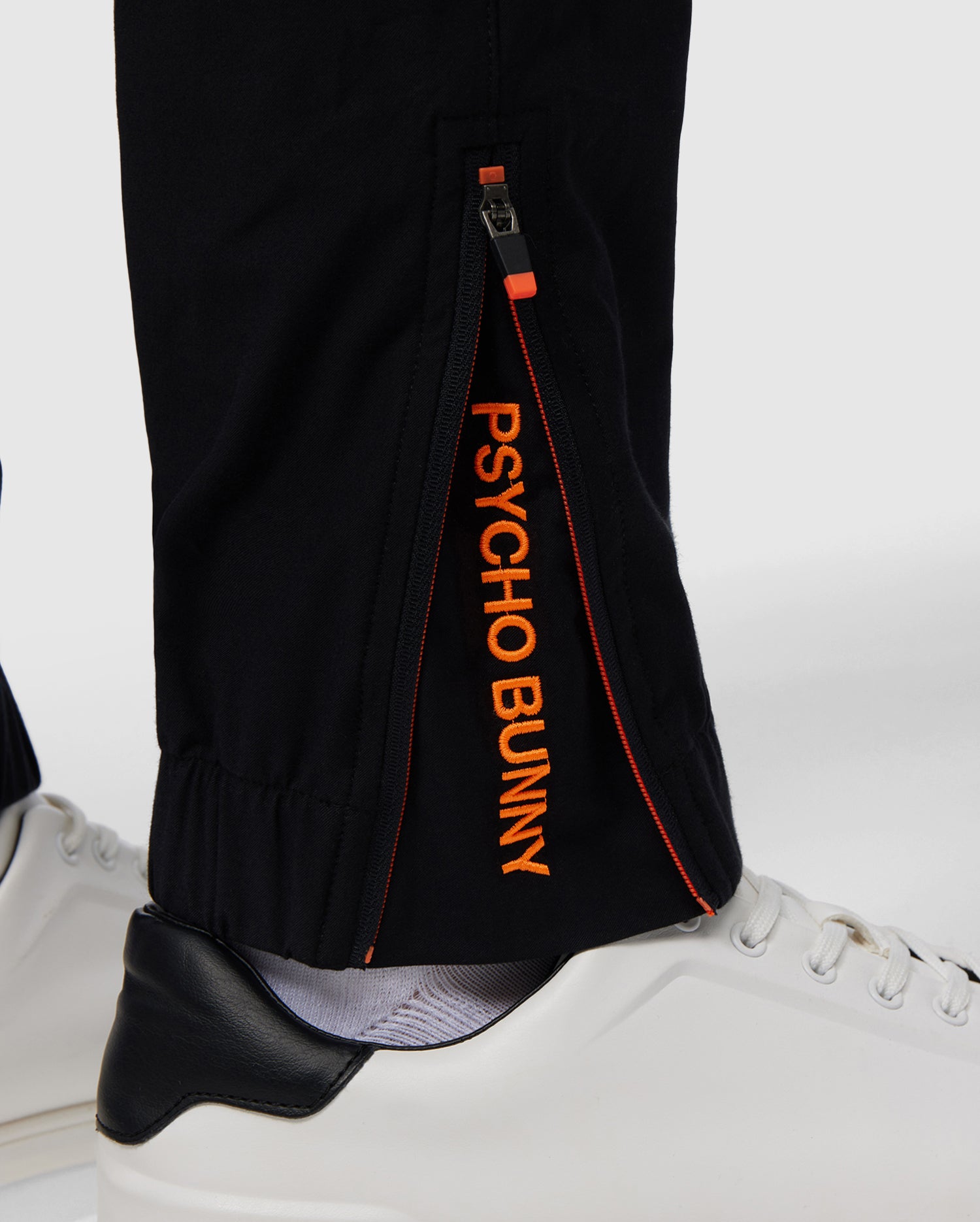 Close-up of the lower leg of black MENS MADISON COMMUTER PANT - B6P395Z1WB by Psycho Bunny, showcasing a zipper detail with the text "psycho bunny" in orange along the seam, paired with white shoes on a white background.