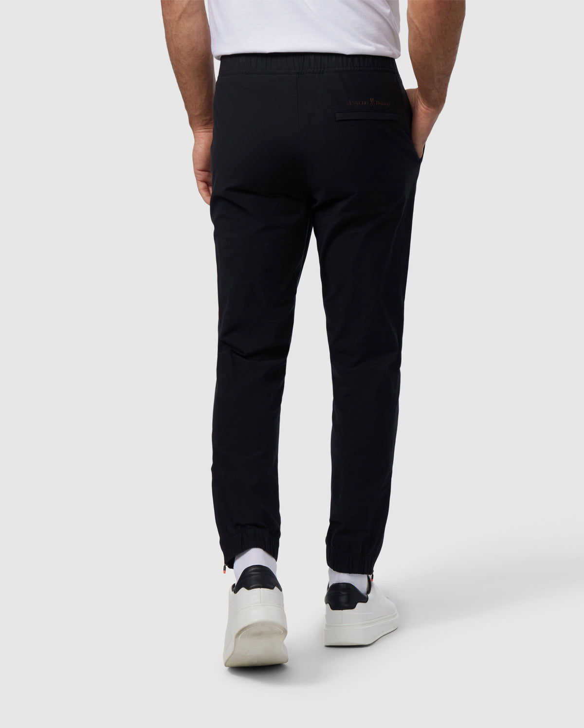 Rear view of a man wearing black Psycho Bunny MENS MADISON COMMUTER PANTS made of stretch woven material and white sneakers, focusing on the lower half of the body against a plain background.