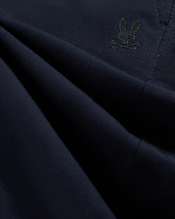 Close-up image of dark blue stretch woven material with subtle folds, featuring a small, embroidered green Psycho Bunny character in the lower left corner.