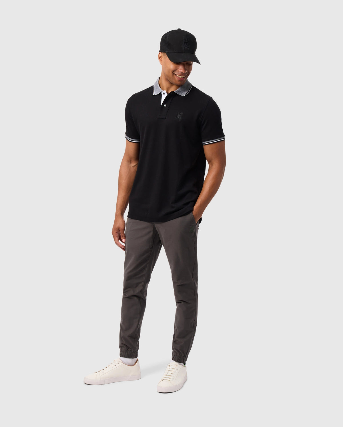 A person dressed in a black polo shirt, wearing Psycho Bunny's MENS MADISON COMMUTER PANT - B6P395Z1WB, which features zippered expansion vents in gray, paired with a black cap and white sneakers stands against a light gray backdrop. They maintain a relaxed posture, smiling while looking slightly downward.