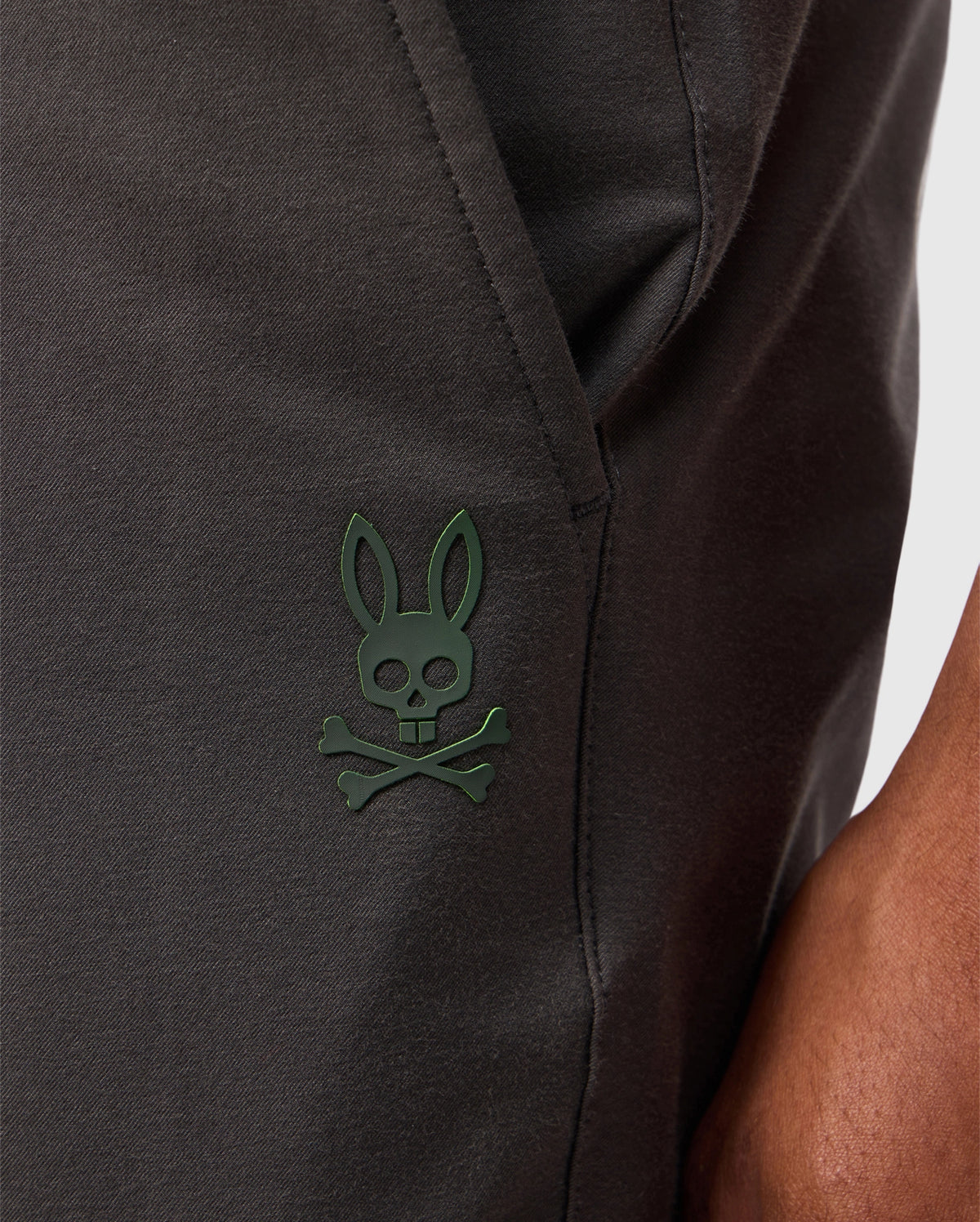 A close-up showcases the Psycho Bunny MENS MADISON COMMUTER PANT - B6P395Z1WB, made from dark fabric with a distinctive green rabbit skull and crossbones emblem. A detachable Bunny keychain dangles nearby, adding character to the design, while zippered expansion vents suggest versatility. A person's arm partially appears on the right.