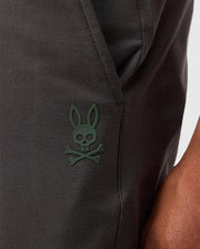 A close-up showcases the Psycho Bunny MENS MADISON COMMUTER PANT - B6P395Z1WB, made from dark fabric with a distinctive green rabbit skull and crossbones emblem. A detachable Bunny keychain dangles nearby, adding character to the design, while zippered expansion vents suggest versatility. A person's arm partially appears on the right.