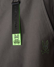 A close-up of the MENS MADISON COMMUTER PANT - B6P395Z1WB by Psycho Bunny reveals a gray fabric with a green tag, featuring their signature black rabbit with crossbones design. Another green emblem displaying the same motif suggests commuter-friendly features like the detachable keychain.