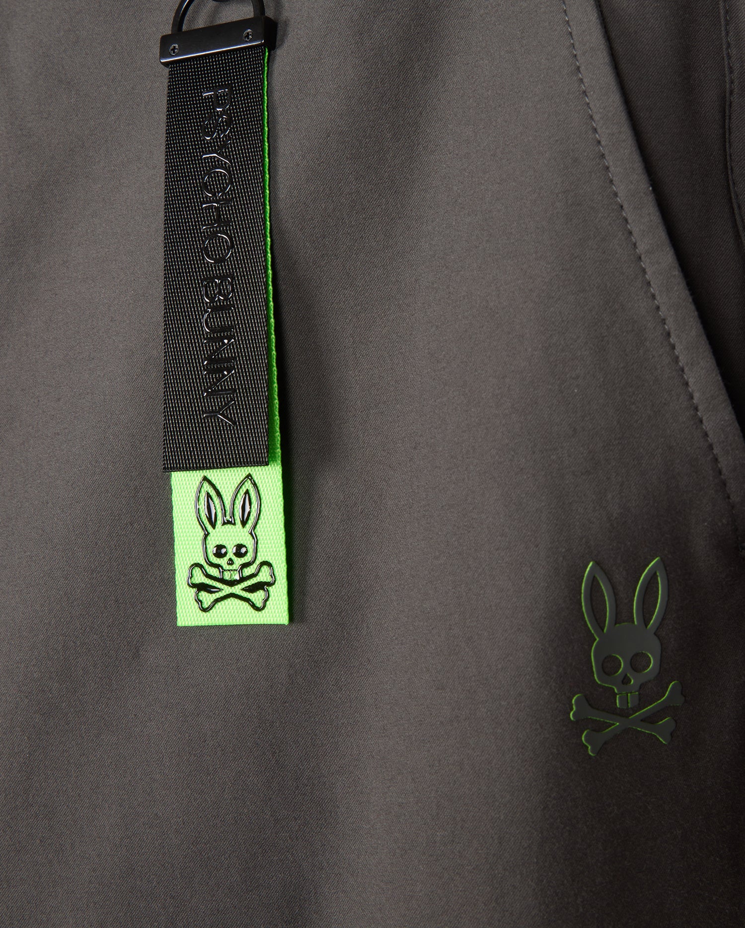 A close-up of the MENS MADISON COMMUTER PANT - B6P395Z1WB by Psycho Bunny reveals a gray fabric with a green tag, featuring their signature black rabbit with crossbones design. Another green emblem displaying the same motif suggests commuter-friendly features like the detachable keychain.