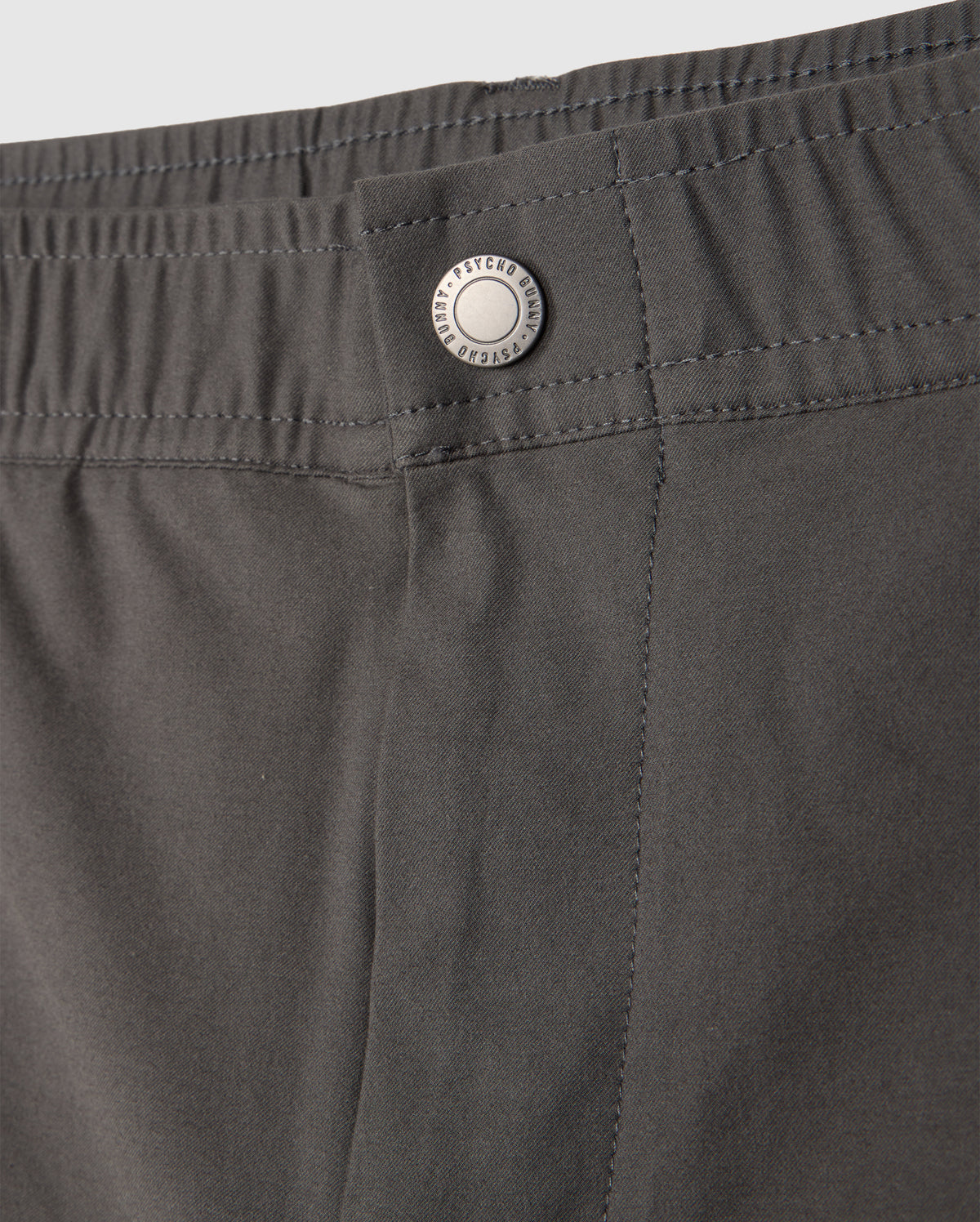 A close-up of the gray MENS MADISON COMMUTER PANT - B6P395Z1WB from Psycho Bunny reveals an elastic waistband and a metal button engraved with "Techwear." The smooth fabric boasts neat stitching and includes a detachable keychain, perfect for the modern commuter.