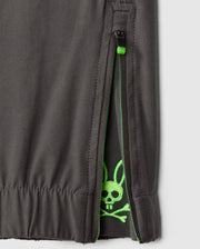 Close-up of the Psycho Bunny MENS MADISON COMMUTER PANT - B6P395Z1WB, featuring a zipper halfway open to reveal a lime green skull and crossbones with rabbit ears design on the inner lining. The dark gray outer fabric pairs well with part of the waistband, and zippered expansion vents provide comfortable wear.