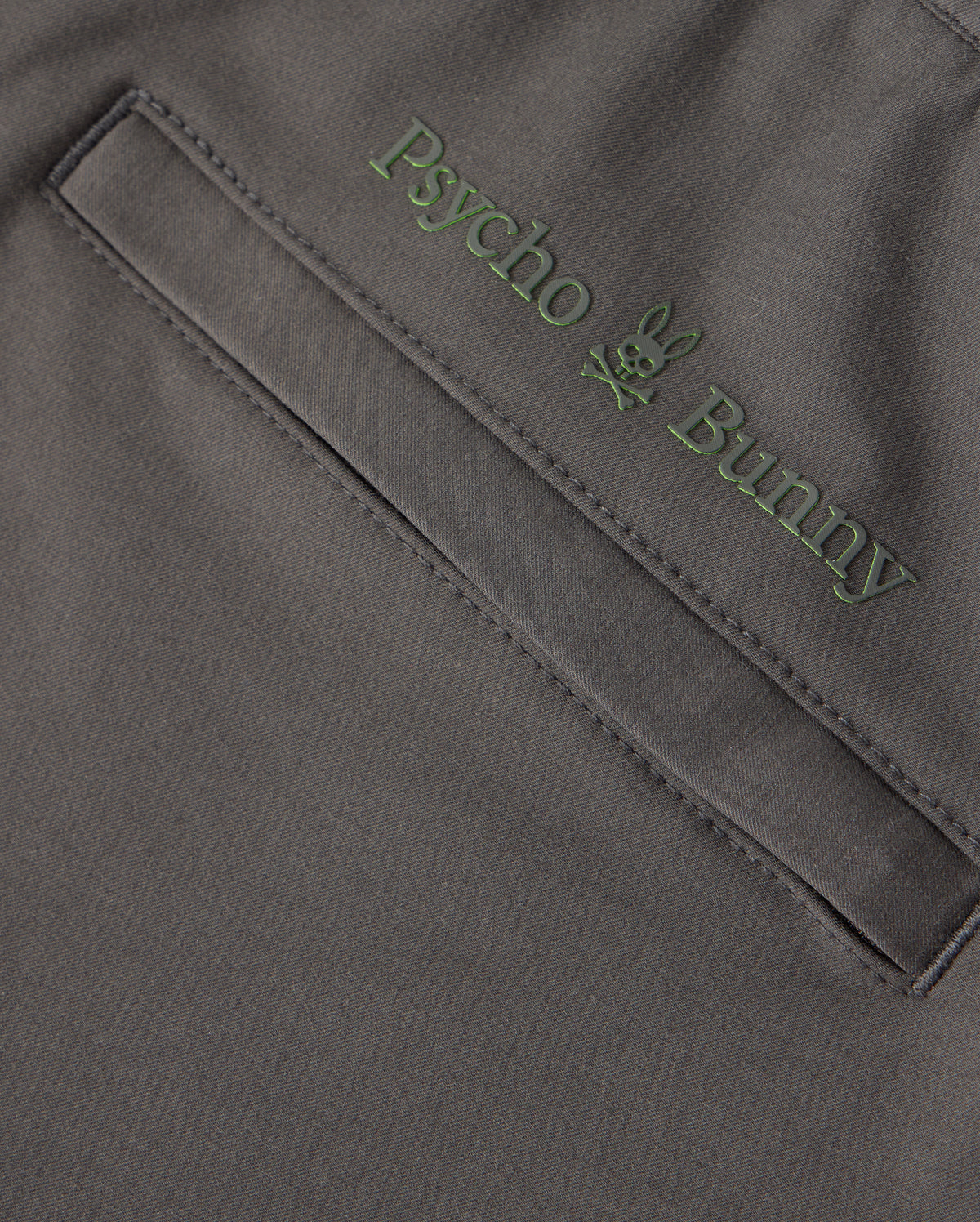 Close-up of the dark gray fabric of the MENS MADISON COMMUTER PANT - B6P395Z1WB, featuring a pocket with green embroidered "Psycho Bunny" text above it. A small bunny logo is positioned between the words, and a detachable keychain enhances both style and functionality.