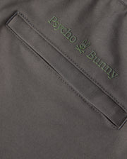 Close-up of the dark gray fabric of the MENS MADISON COMMUTER PANT - B6P395Z1WB, featuring a pocket with green embroidered "Psycho Bunny" text above it. A small bunny logo is positioned between the words, and a detachable keychain enhances both style and functionality.