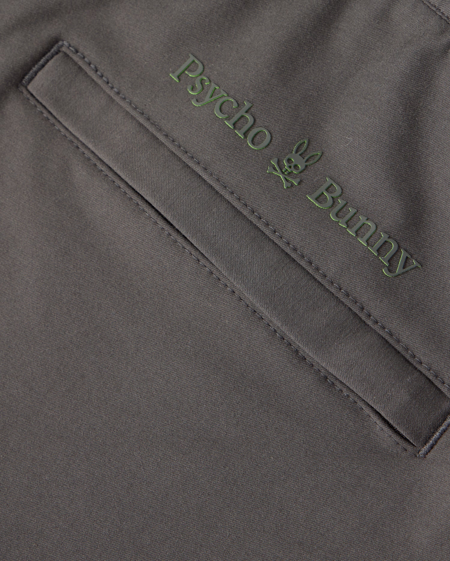 Close-up of the dark gray fabric of the MENS MADISON COMMUTER PANT - B6P395Z1WB, featuring a pocket with green embroidered "Psycho Bunny" text above it. A small bunny logo is positioned between the words, and a detachable keychain enhances both style and functionality.