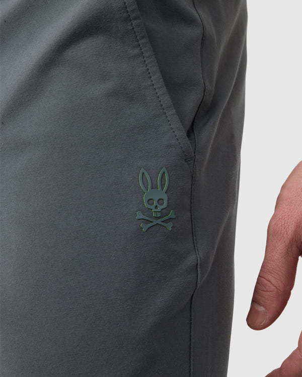 Close-up of a dark gray fabric with a small green skull and crossbones logo, depicting a character with bunny ears, embroidered near the elasticized waistband on the Psycho Bunny MENS MADISON COMMUTER PANT - B6P395Z1WB.