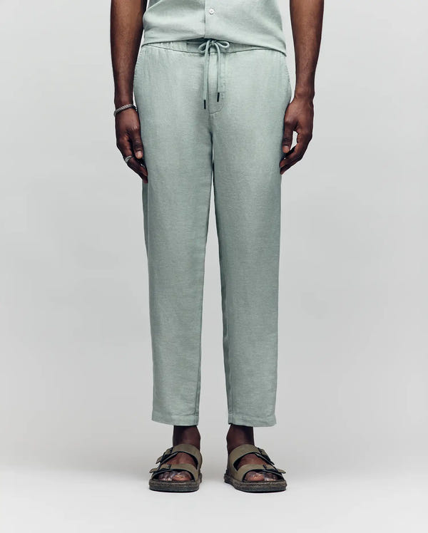 A person stands facing forward, wearing Psycho Bunny MENS FATE LINEN DRAWSTRING PANT (B6P471C200) in light sage green with brown sandals. Their hands are at their sides, ideal for summer, against a plain, light backdrop.