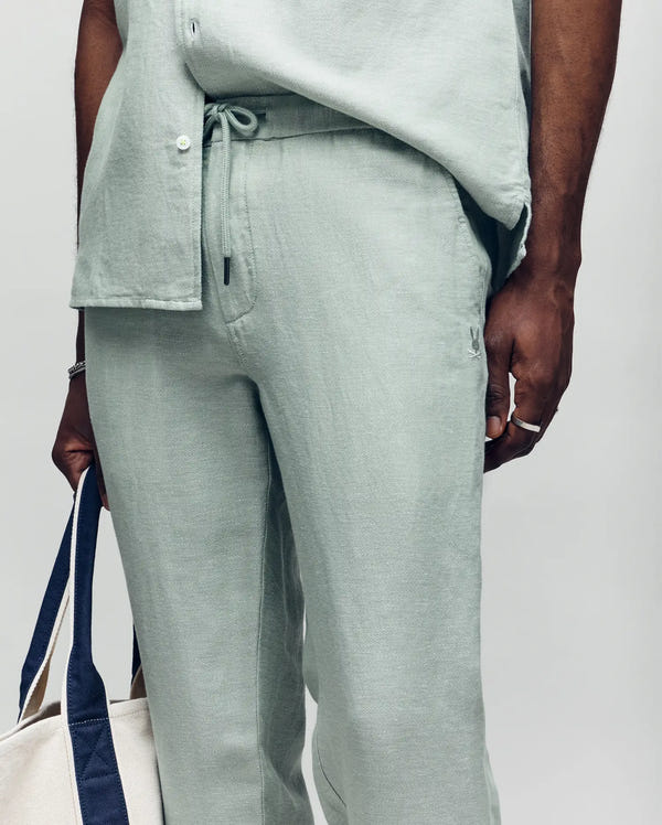 A person wears Psycho Bunny’s MENS FATE LINEN DRAWSTRING PANT in light green with a matching shirt, holding a white tote bag with navy blue handles. Only the lower torso and hand are visible, highlighting effortless summer style.