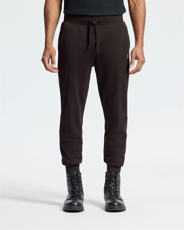 A person in Psycho Bunny's MENS LEVY ESSENTIAL FLEECE SWEATPANT (B6P721D200) and black lace-up boots stands against a white background, exuding a relaxed vibe with their arms at their sides.
