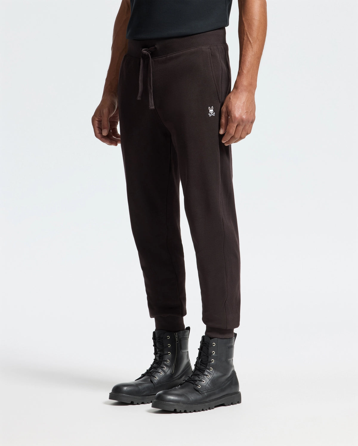The lower half of the person wears Psycho Bunny's MENS LEVY ESSENTIAL FLEECE SWEATPANT in black, complete with a small embroidered logo and drawstring waist. Paired with black lace-up boots against a light background, these jogger pants offer a perfect casualwear look.