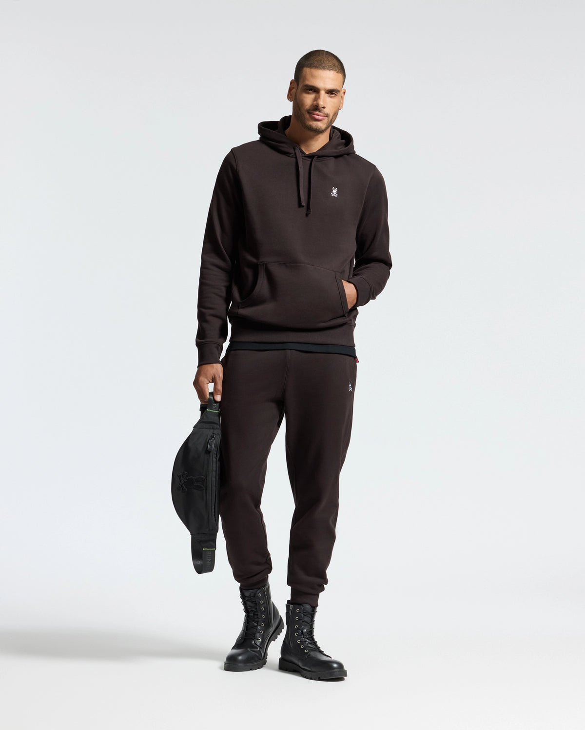 A person in a black hoodie and Psycho Bunny's MENS LEVY ESSENTIAL FLEECE SWEATPANT (B6P721D200) holds a black bag. They wear black boots, standing against a plain light background.