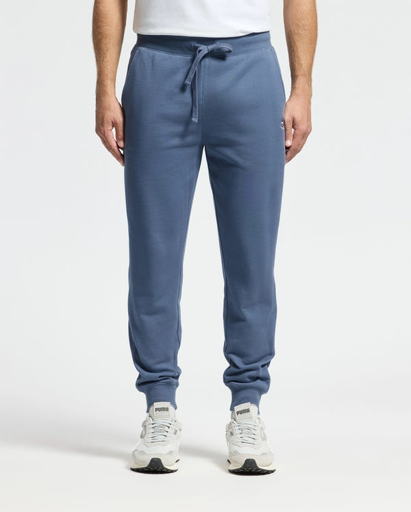 A person is wearing comfortable blue Psycho Bunny MEN'S LEVY ESSENTIAL FLEECE SWEATPANTS - B6P721D200 and white sneakers. Their upper body is partially visible, revealing a white t-shirt against a plain white background.