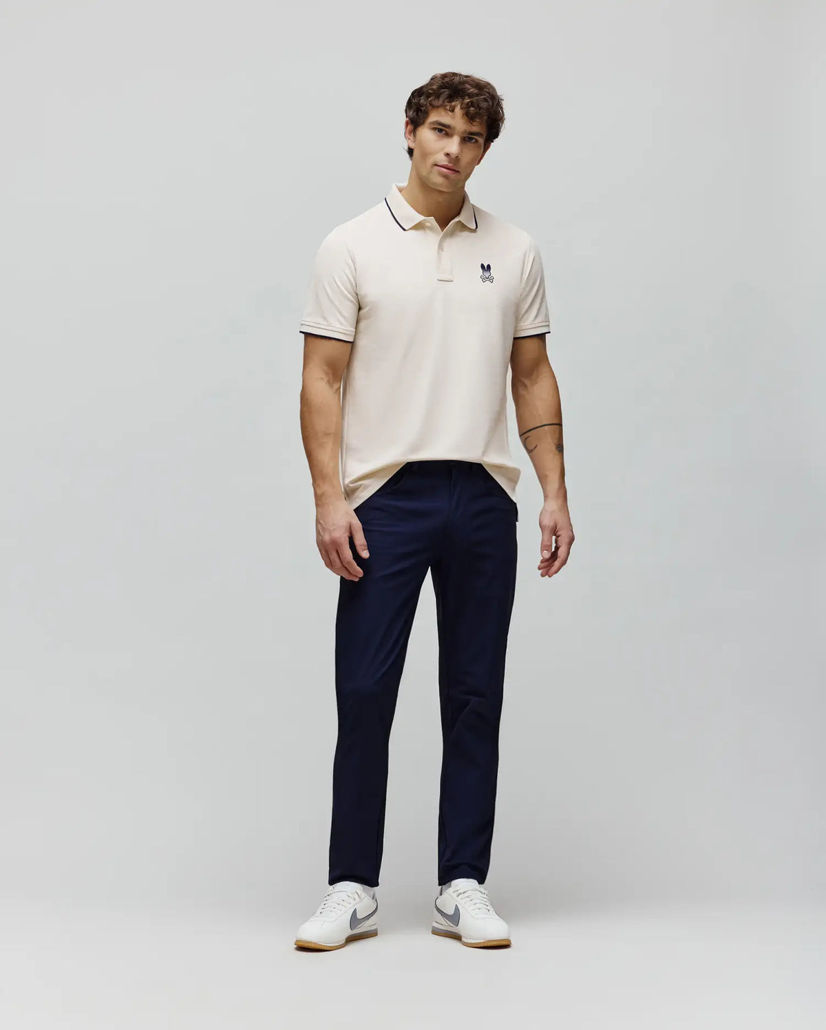 Classic fit cream pique polo with navy tipping and small embroidered Psycho Bunny skull logo on left chest, paired with sleek navy Mens Maverick 10K Step Pant.