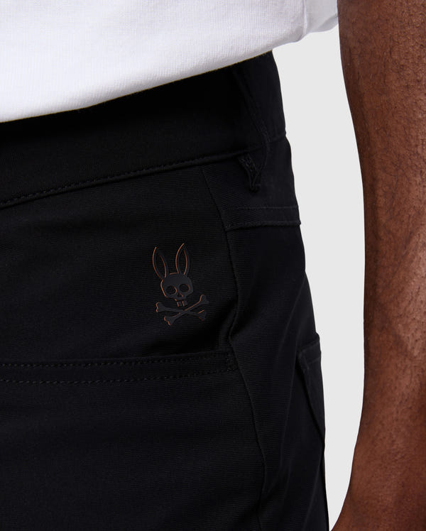 A close-up view showcases someone wearing Psycho Bunny's MENS MAVERICK 10K PANT - B6P788D200 in black, highlighted by the distinctive skull and bunny ears logo on the pocket. These four-way stretch pants offer a blend of comfort and style, impeccably paired with their crisp white shirt.