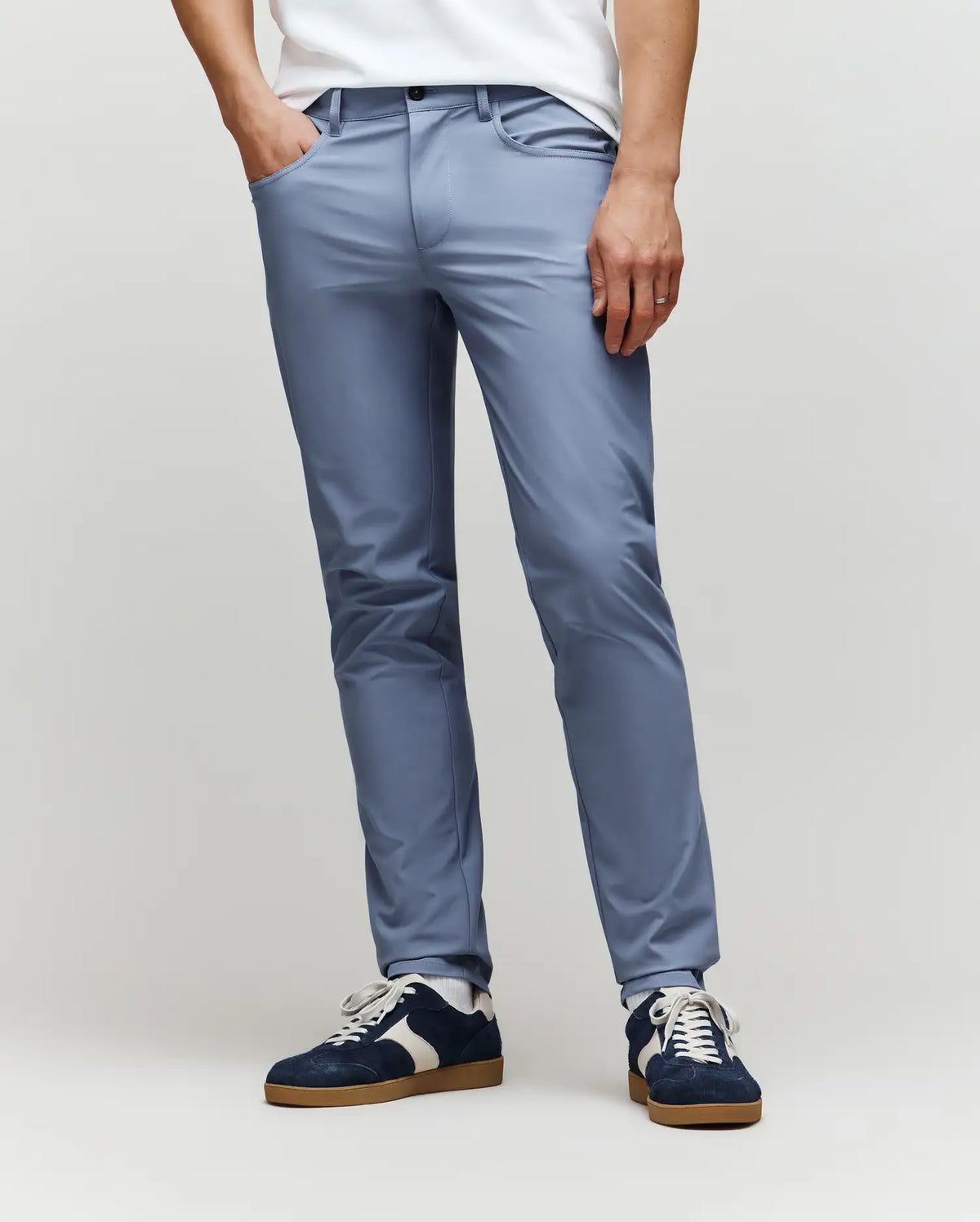 A person, visible from the waist down, stands against a plain light gray background wearing Psycho Bunny MENS MAVERICK 10K STEP PANT - B6P788D200 in light blue with five-pocket styling, paired with a white shirt and navy sneakers with white laces.