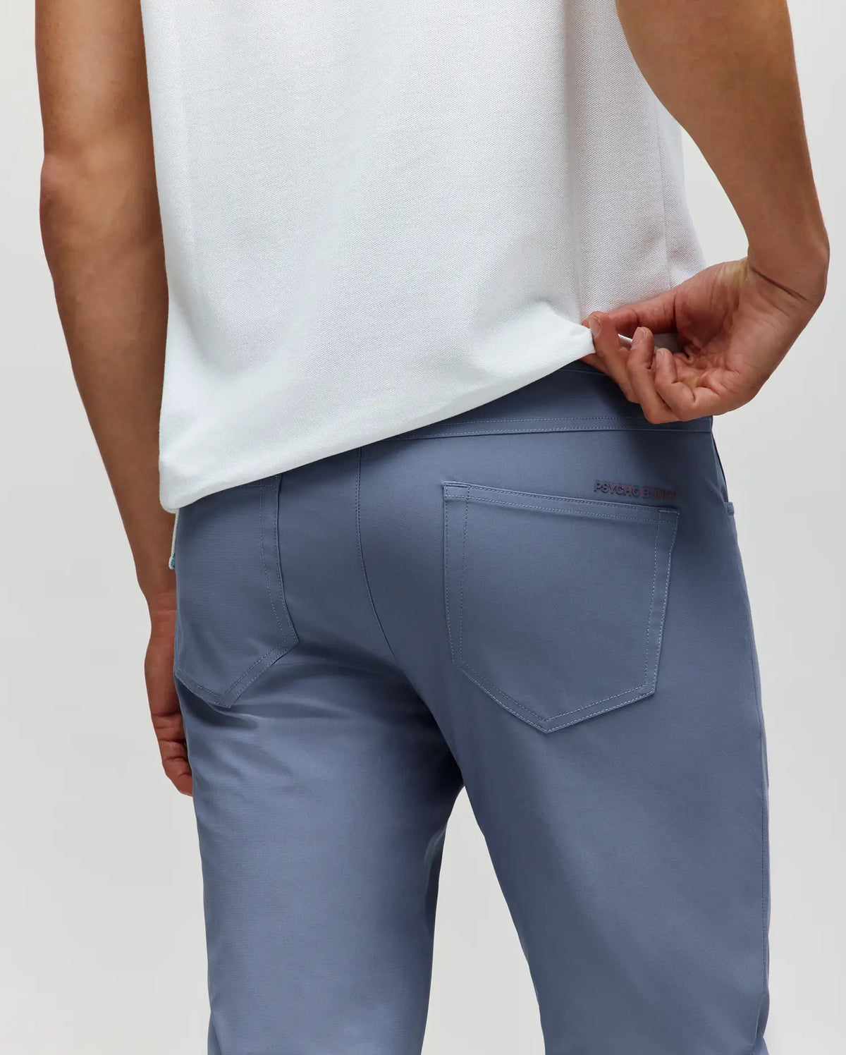 A person in light blue Psycho Bunny MENS MAVERICK 10K STEP PANT (B6P788D200) with five-pocket styling wears a white shirt, lifting it to show the rear pockets, photographed from behind against a plain background.