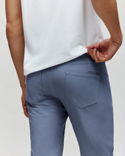 A person in light blue Psycho Bunny MENS MAVERICK 10K STEP PANT (B6P788D200) with five-pocket styling wears a white shirt, lifting it to show the rear pockets, photographed from behind against a plain background.