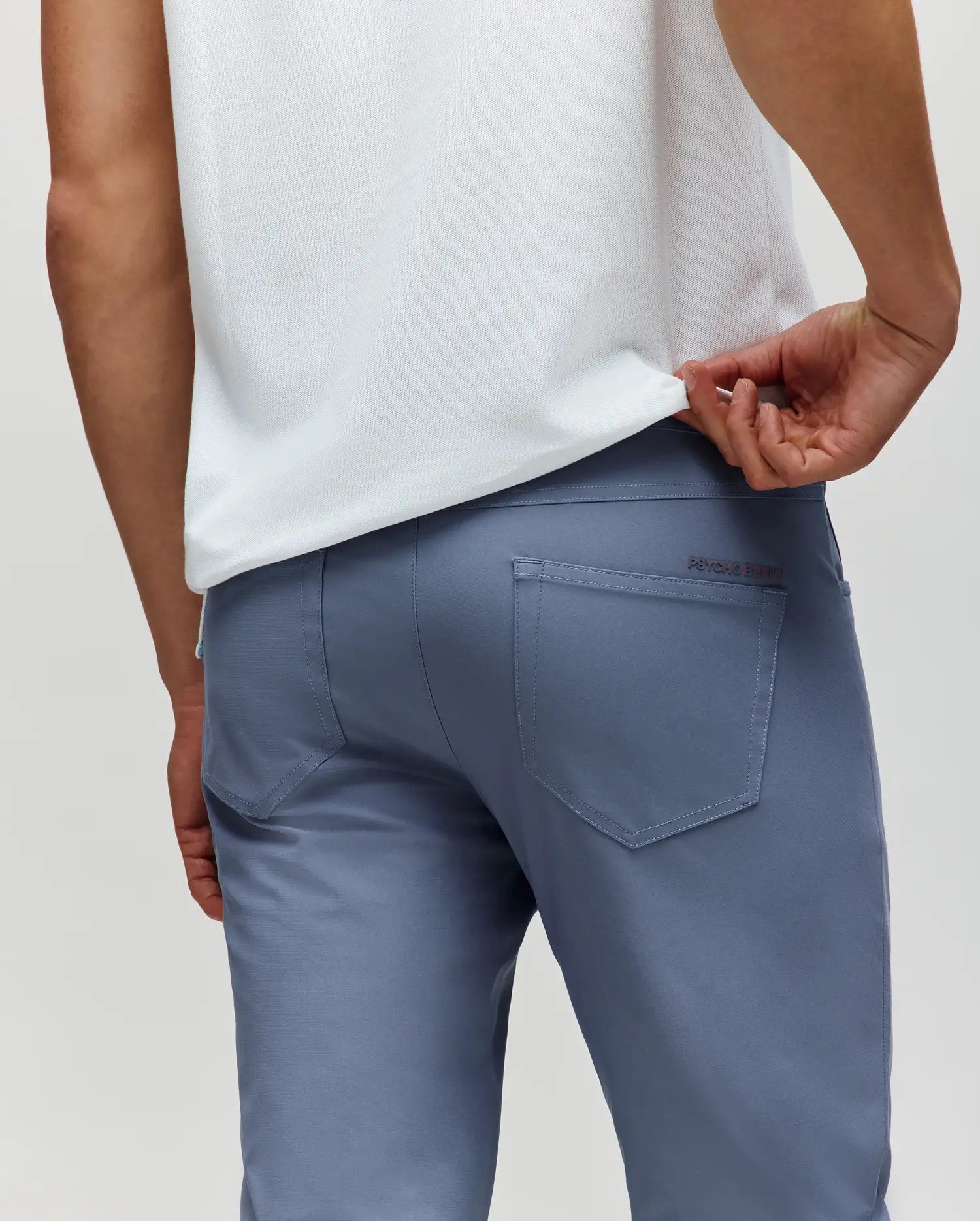 A person in light blue Psycho Bunny MENS MAVERICK 10K STEP PANT (B6P788D200) with five-pocket styling wears a white shirt, lifting it to show the rear pockets, photographed from behind against a plain background.
