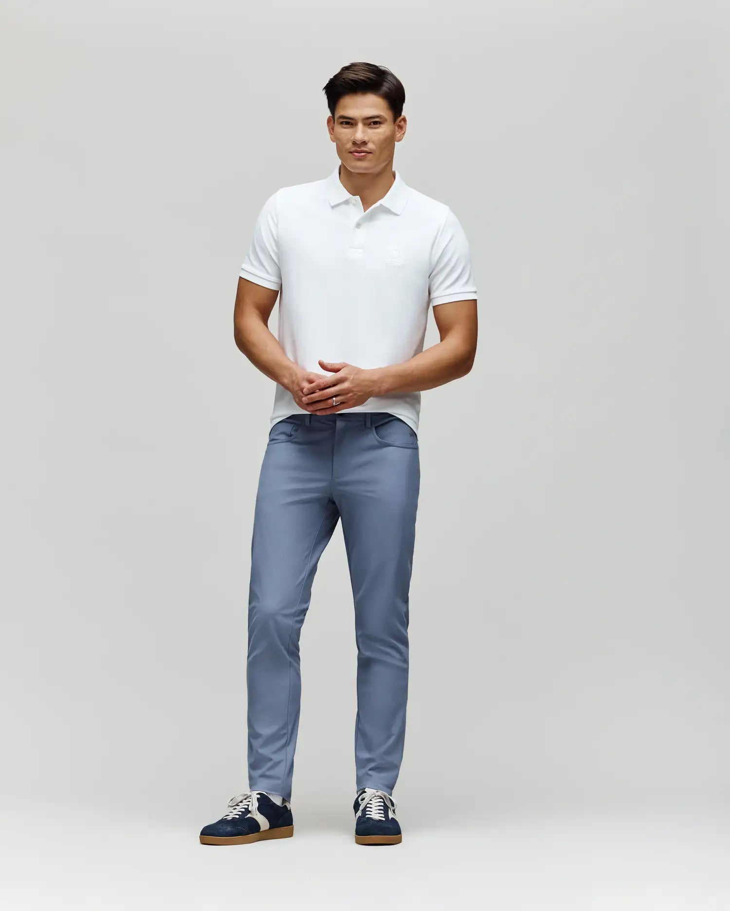 A man stands against a light gray background, wearing a white short-sleeve polo and Psycho Bunny MENS MAVERICK 10K STEP PANT - B6P788D200 in light blue with four-way stretch and five-pocket styling. He faces forward, hands clasped.