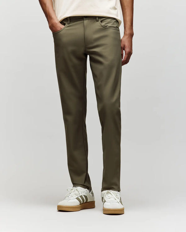 A person in Psycho Bunny’s MENS MAVERICK 10K STEP PANT - B6P788D200 (olive green, five-pocket styling), a cream t-shirt, and white sneakers with gray stripes stands against a light gray backdrop; upper body not fully visible.