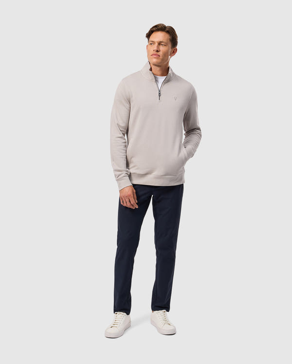 A man stands against a plain background wearing a light gray quarter-zip sweater, Psycho Bunny's MENS MAVERICK 10K PANT in dark navy (B6P788D200), and white sneakers. His hands are in his pockets, glancing to the side.