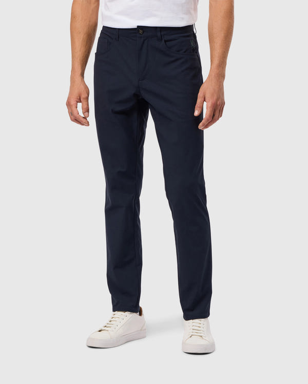 A person wearing dark blue MENS MAVERICK 10K PANT - B6P788D200 by Psycho Bunny and white sneakers stands against a light gray background, with the pants featuring a single button, paired with a tucked-in white shirt.