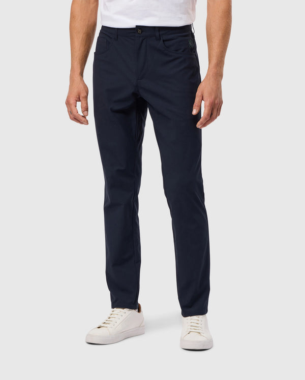 A person wearing dark blue MENS MAVERICK 10K PANT - B6P788D200 by Psycho Bunny and white sneakers stands against a light gray background, with the pants featuring a single button, paired with a tucked-in white shirt.