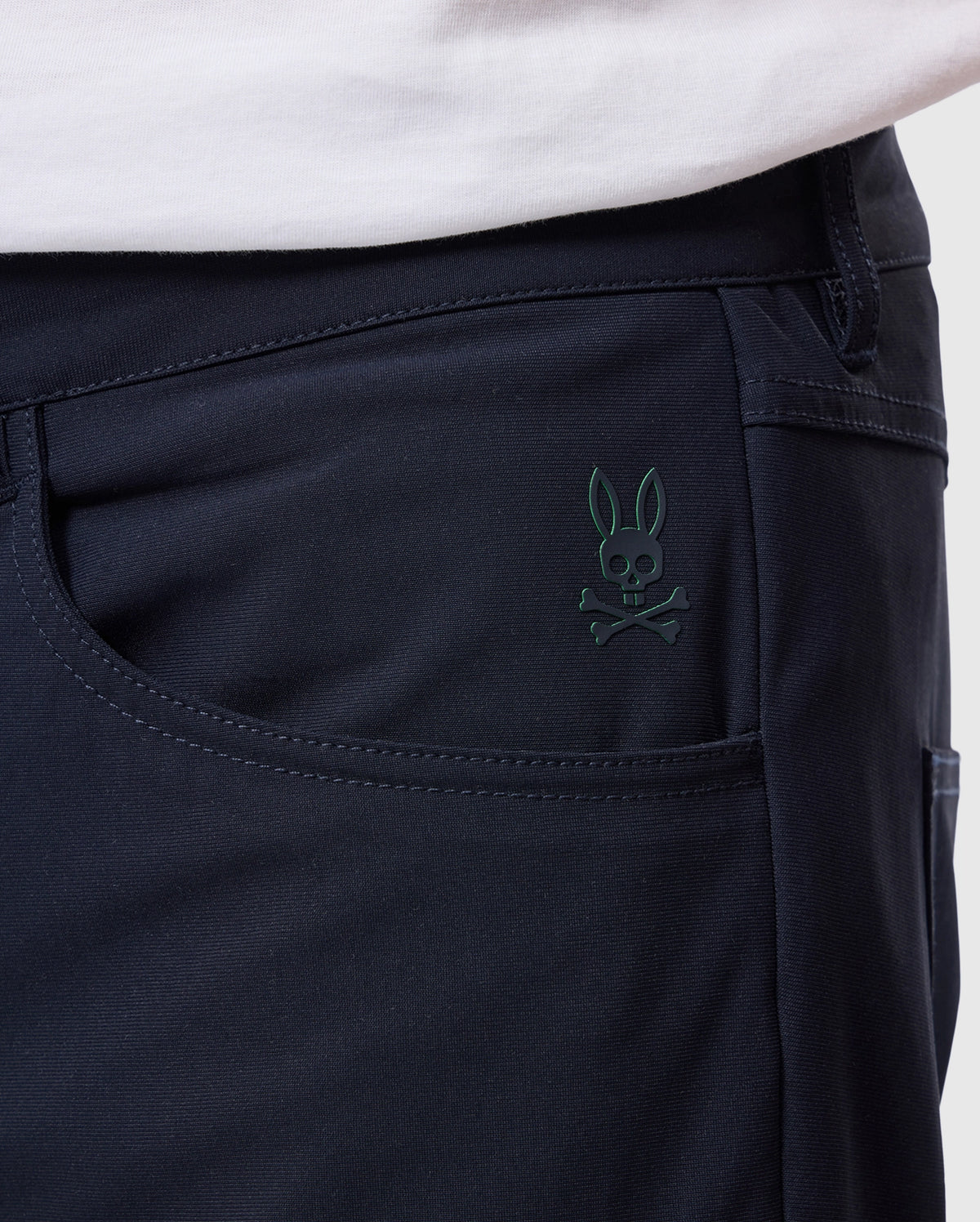Close-up of someone wearing Psycho Bunny's "MENS MAVERICK 10K PANT" displaying a subtle skull and bunny ears logo on the black fabric pocket, with part of a white shirt visible above.