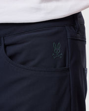 Close-up of someone wearing Psycho Bunny's "MENS MAVERICK 10K PANT" displaying a subtle skull and bunny ears logo on the black fabric pocket, with part of a white shirt visible above.
