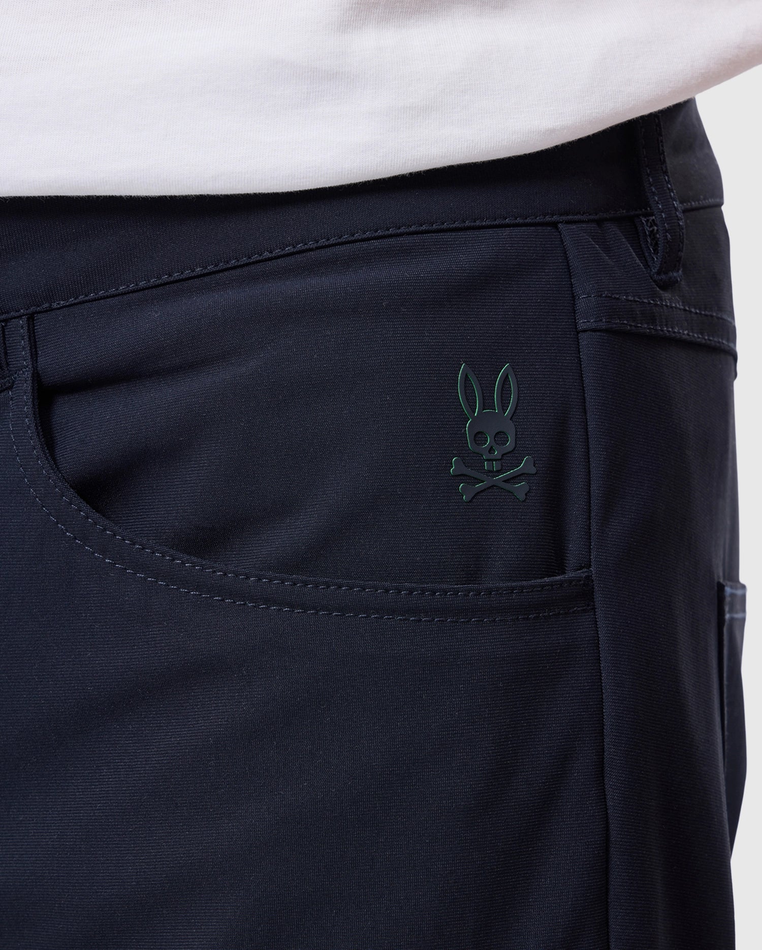 Close-up of someone wearing Psycho Bunny's "MENS MAVERICK 10K PANT" displaying a subtle skull and bunny ears logo on the black fabric pocket, with part of a white shirt visible above.