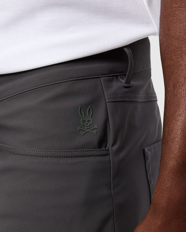 Close-up of a person wearing Psycho Bunny's MENS 10K STEP PANT - B6P788D200 in dark gray, featuring a five-pocket design with an additional small side pocket that bears the stitched emblem of a bunny skull and crossbones. The person is also seen wearing a white textured shirt, with their hand resting near the pocket.