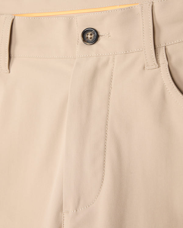 A close-up view of the Psycho Bunny MENS 10K STEP PANT - B6P788D200 beige four-way stretch trousers focusing on the waistband, a black button, and belt loops. The fabric appears smooth and well-stitched, showcasing the front pocket detail and a part of the yellow lining inside the waistband.