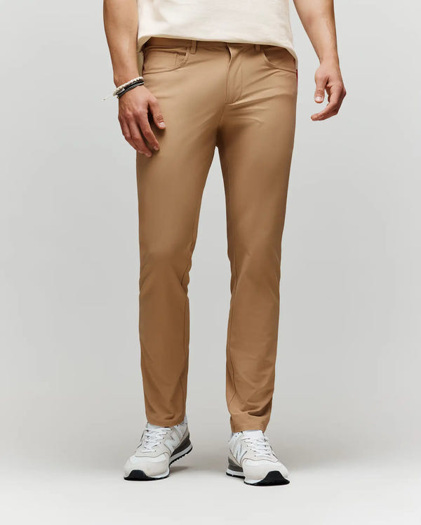 A person in tan MENS MAVERICK 10K STEP PANT - B6P788D200 by Psycho Bunny, paired with a light shirt, white sneakers, and bracelets on their left wrist, stands against a plain light background.