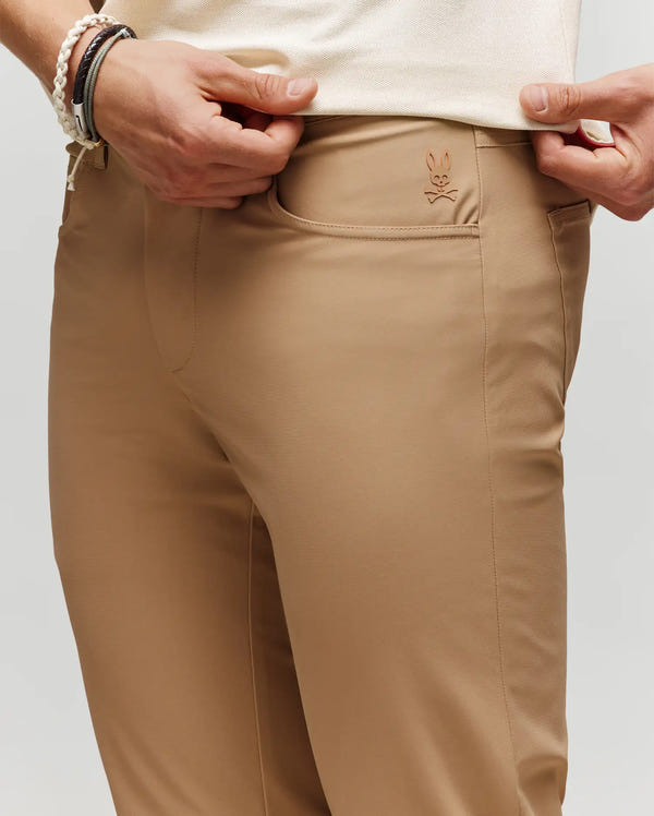 A person wearing tan Psycho Bunny MENS MAVERICK 10K STEP PANT (B6P788D200) with a small bunny logo above the front pocket lifts their shirt to display the pants, while several bracelets adorn their wrist.