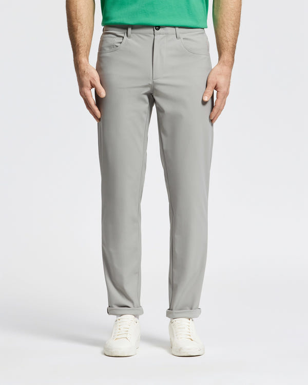 A person is wearing the light gray Psycho Bunny MENS MAVERICK 10K STEP PANT - B6P788D200 featuring five-pocket styling paired with a green shirt and white sneakers, standing with hands by their sides. The backdrop is plain and light-colored.
