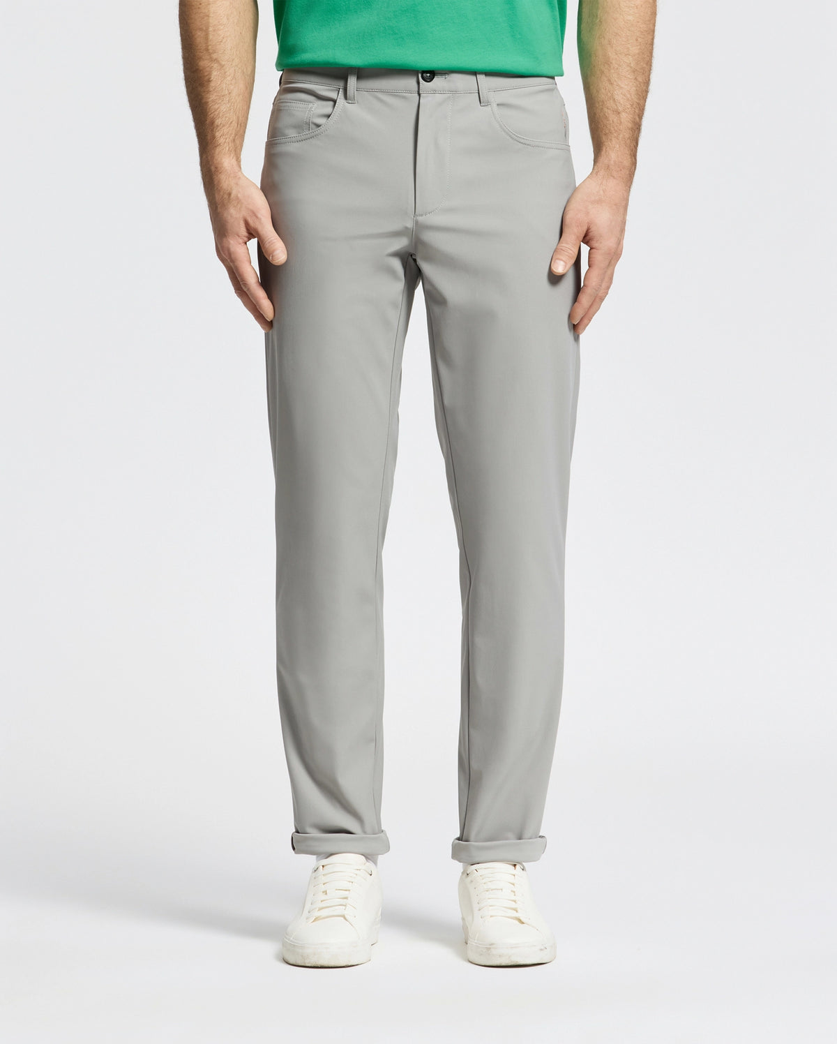 A person is wearing the light gray Psycho Bunny MENS MAVERICK 10K STEP PANT - B6P788D200 featuring five-pocket styling paired with a green shirt and white sneakers, standing with hands by their sides. The backdrop is plain and light-colored.