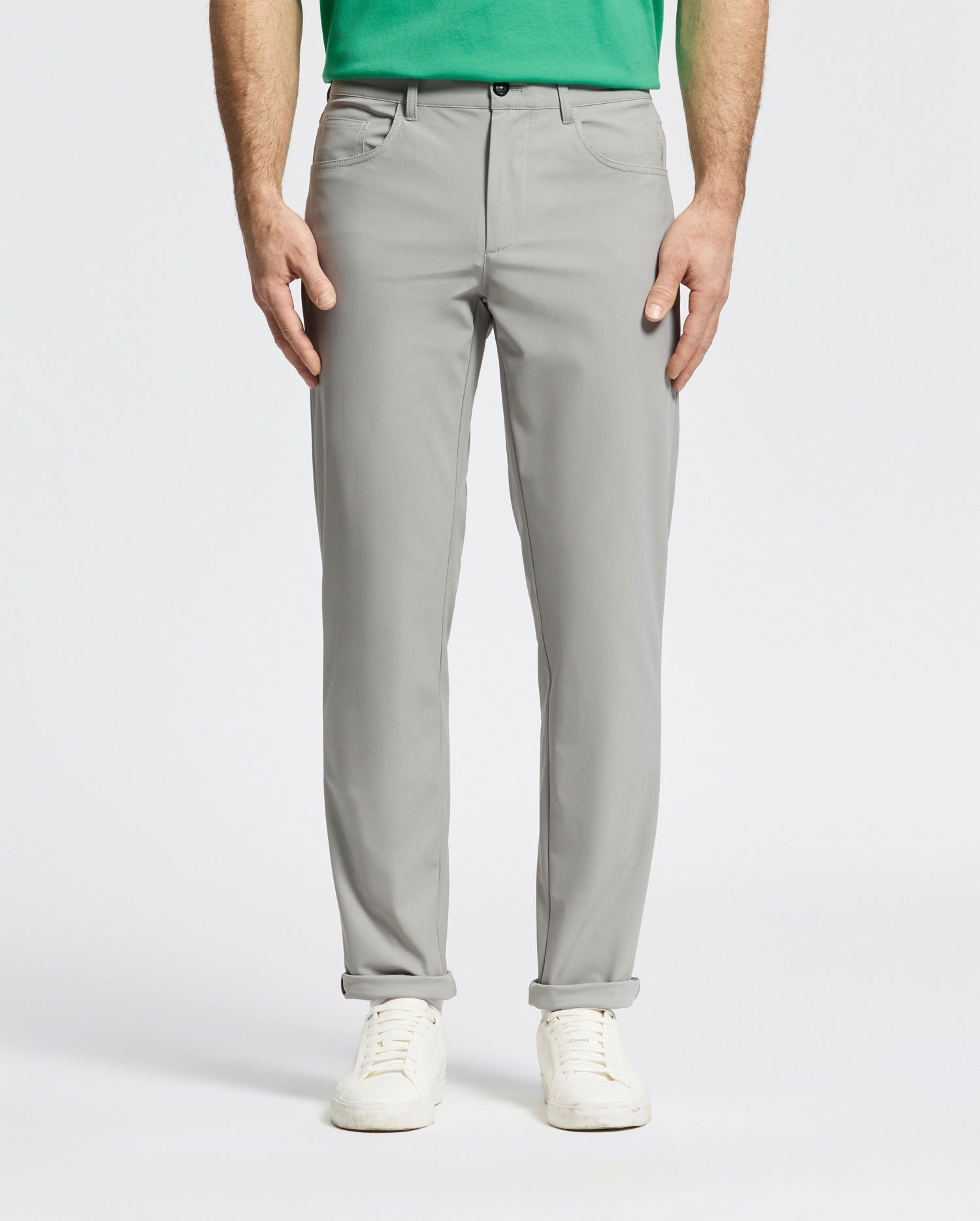 A person is wearing the light gray Psycho Bunny MENS MAVERICK 10K STEP PANT - B6P788D200 featuring five-pocket styling paired with a green shirt and white sneakers, standing with hands by their sides. The backdrop is plain and light-colored.