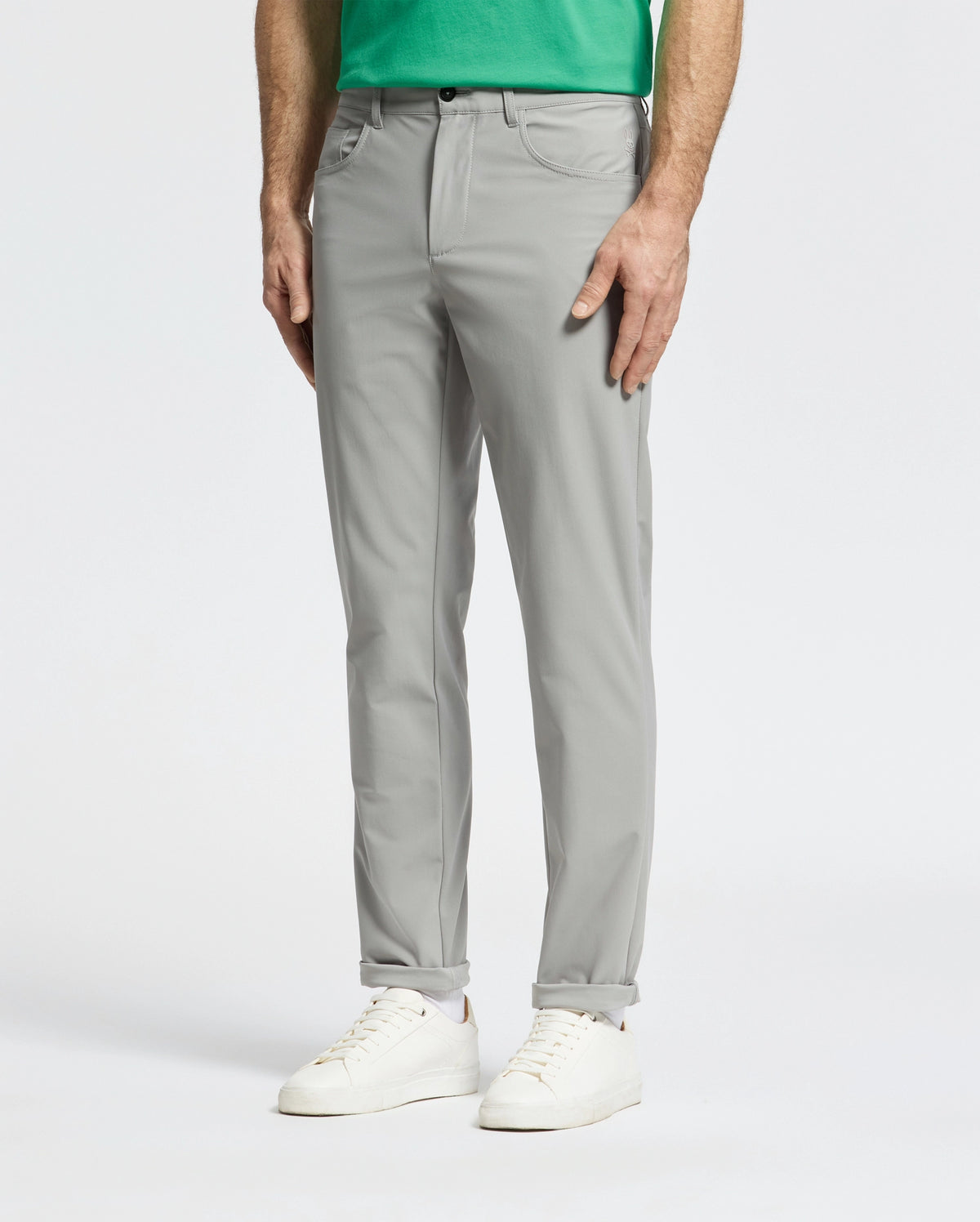 A person models the Psycho Bunny MENS MAVERICK 10K STEP PANT - B6P788D200 in light gray with five pockets, paired with a green shirt and white sneakers, highlighting comfort and style on a plain white background.