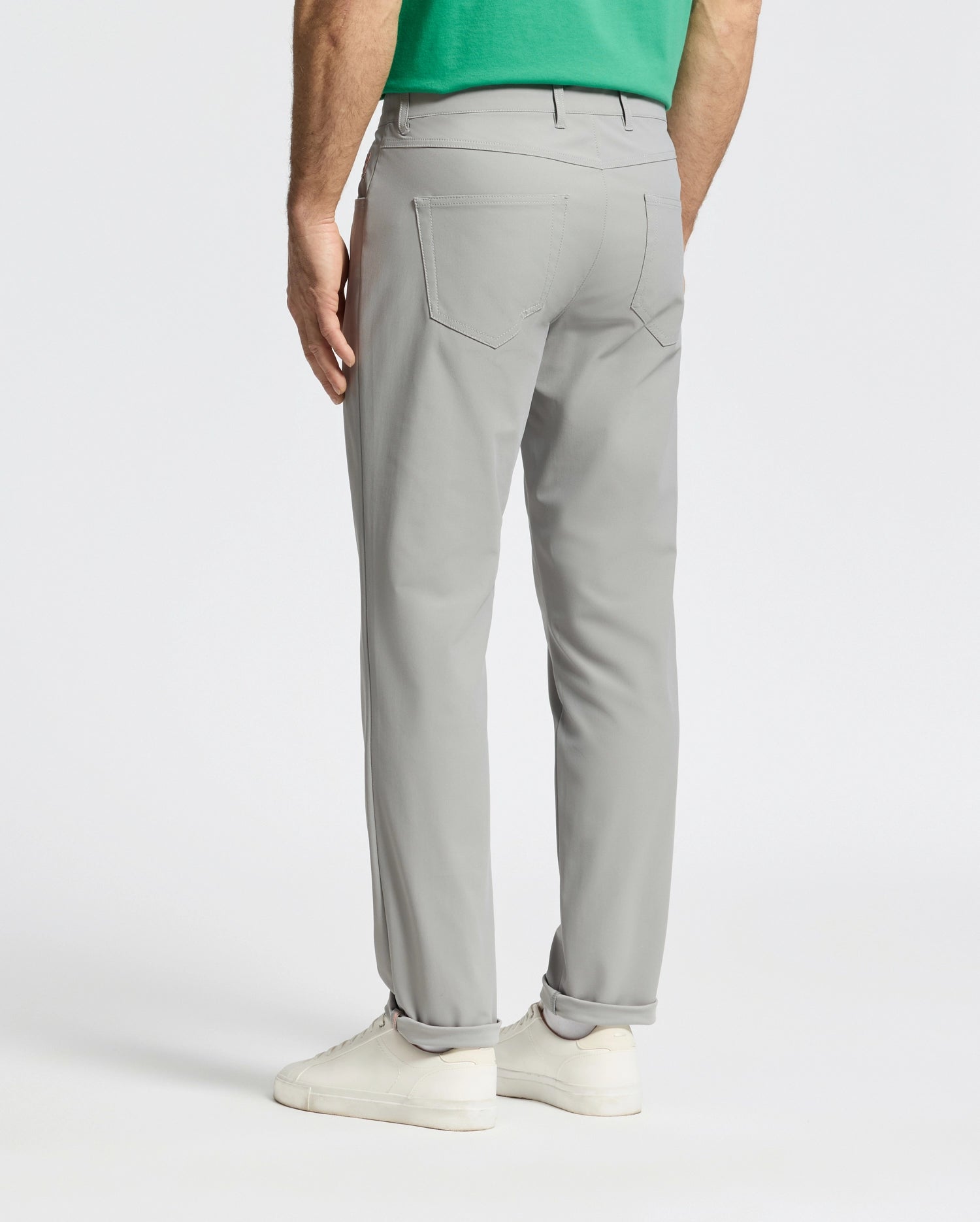 Wearing Psycho Bunny's MENS MAVERICK 10K STEP PANT in light gray, paired with white sneakers and a green shirt, a person stands facing away. The plain background suggests a studio setting.