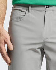 A person wears Psycho Bunny's MENS MAVERICK 10K STEP PANT - B6P788D200 in light gray with four-way stretch and a black button, paired with a green shirt. Their left hand rests near the pocket as the focus is on the pants and part of the shirt against a white background.