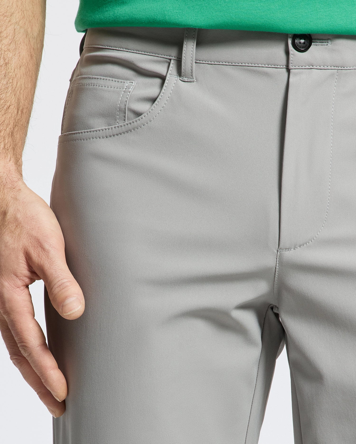 A person wears Psycho Bunny's MENS MAVERICK 10K STEP PANT - B6P788D200 in light gray with four-way stretch and a black button, paired with a green shirt. Their left hand rests near the pocket as the focus is on the pants and part of the shirt against a white background.