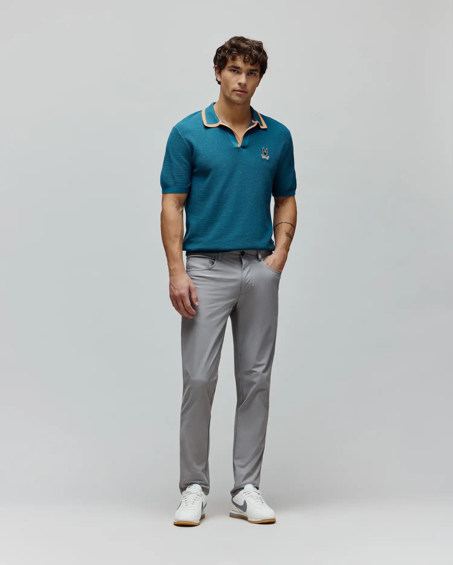 Teal knit polo with orange collar trim and small embroidered Psycho Bunny logo on left chest, paired with slim fit light grey Mens Maverick 10k Step Pant.