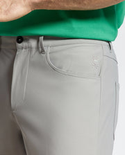 A person wears MENS MAVERICK 10K STEP PANT - B6P788D200 in light gray with an embroidered bunny above crossed bones on the pocket. Paired with a green shirt, it showcases Psycho Bunny's five-pocket styling and four-way stretch, emphasizing side and front pocket details.