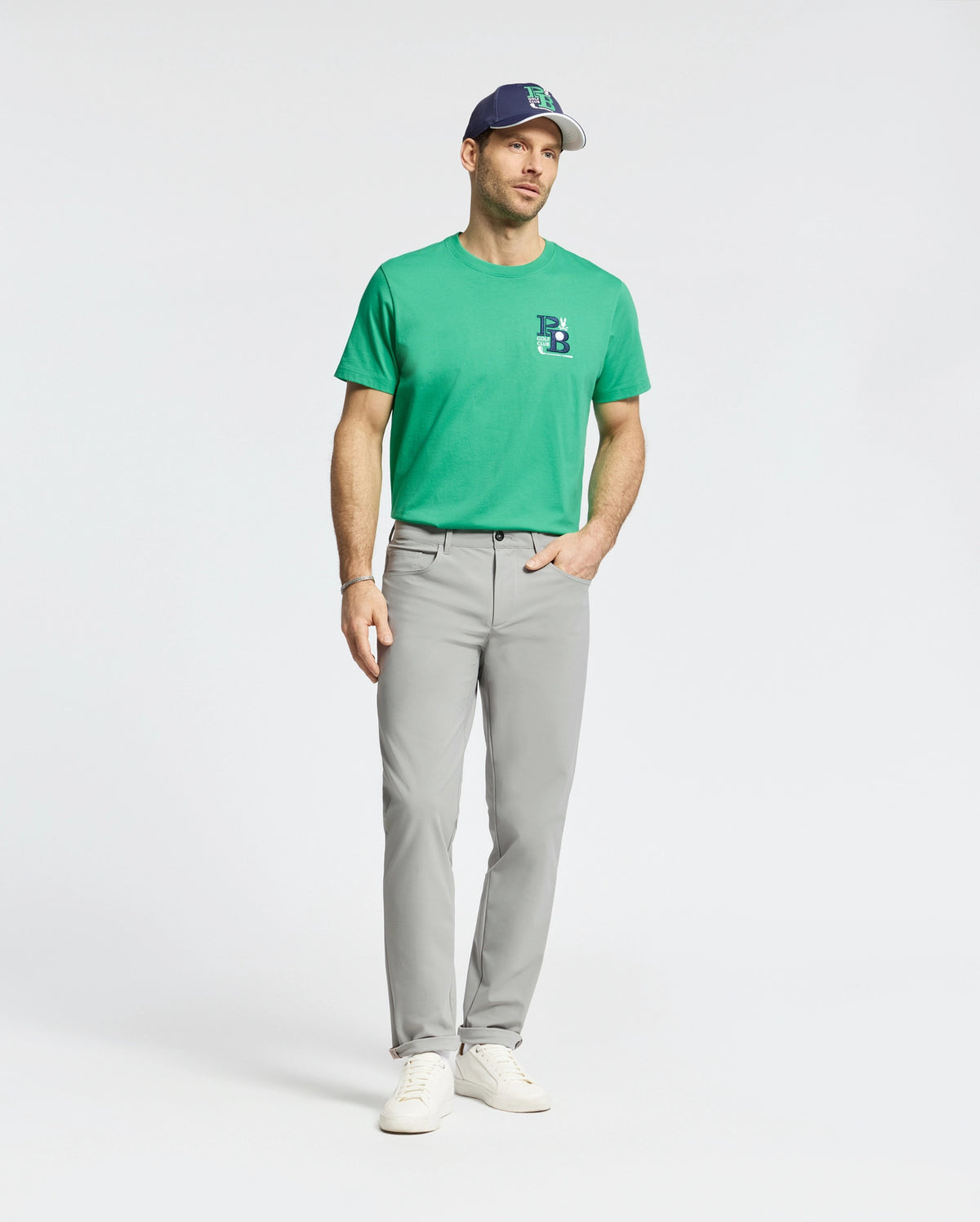 A man dons a green T-shirt with a subtle logo, the Psycho Bunny MENS MAVERICK 10K STEP PANT B6P788D200 in light gray featuring five-pocket styling, white sneakers, and a blue and white baseball cap. He stands confidently against a plain white background.