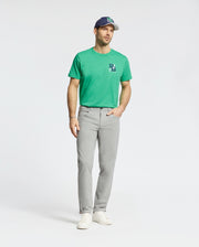 A man dons a green T-shirt with a subtle logo, the Psycho Bunny MENS MAVERICK 10K STEP PANT B6P788D200 in light gray featuring five-pocket styling, white sneakers, and a blue and white baseball cap. He stands confidently against a plain white background.